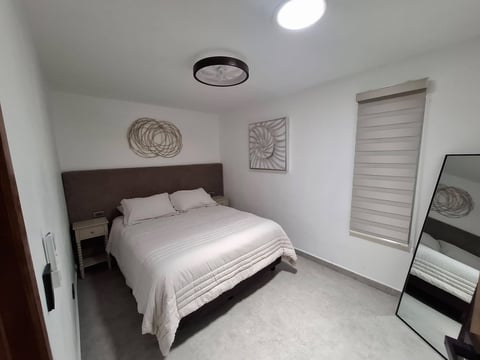 Recámara con cabecera café, cama queen, ropa de cama blanca, arte circular en pared, persianas enrollables, espejo de piso y foco de techo