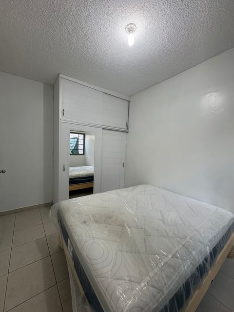 Recámara minimalista con colchón envuelto en plástico, clóset empotrado blanco, piso de loseta y foco de techo