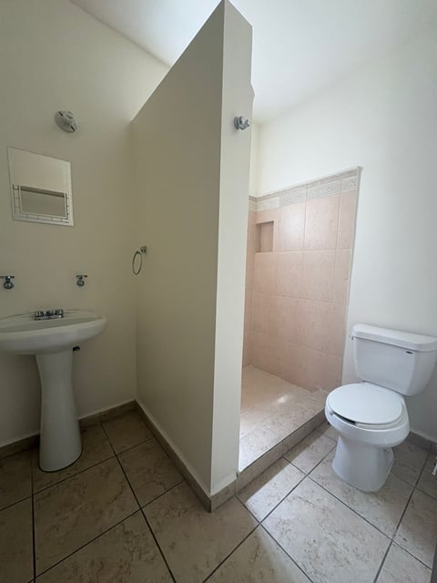 Baño con lavamanos tipo pedestal, inodoro blanco, área de regadera con azulejo rosa y piso cerámico beige