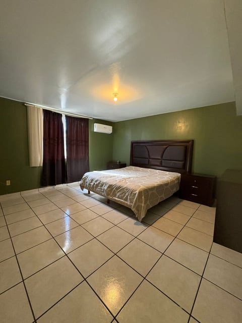 Recámara principal con paredes verdes, cama beige con edredón dorado y cortinas oscuras