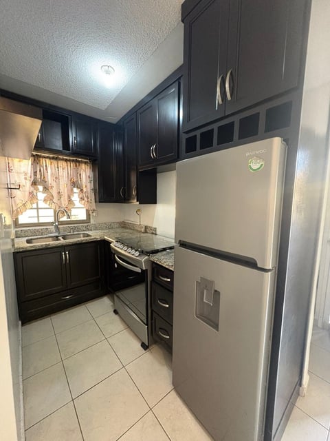 Cocina pequeña con gabinetes negros, refrigerador color beige, estufa, tarja y piso de loseta con ventana y cortinas