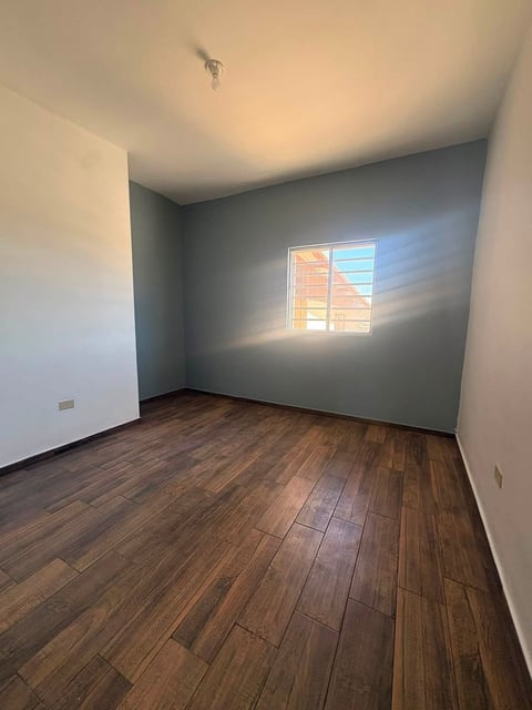 Recámara vacía con piso de madera, paredes blancas y gris, y luz natural desde ventana