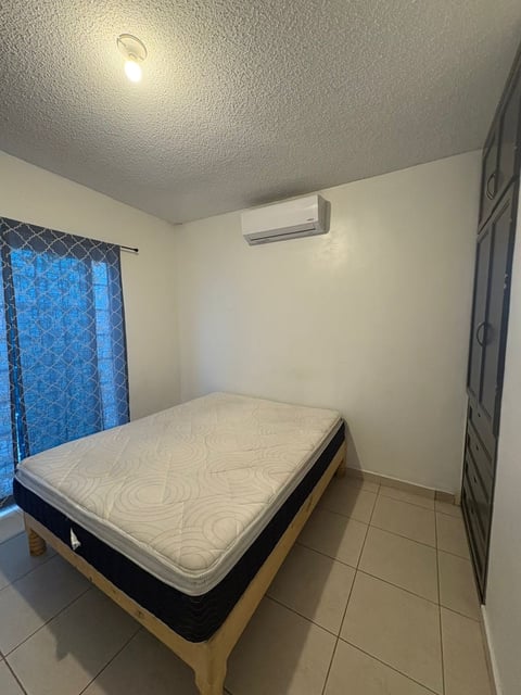 Recámara minimalista con cama, minisplit en pared, iluminación de techo y cortinas azules con patrón sobre piso de loseta