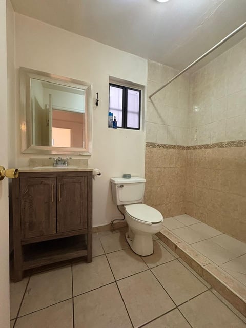 Baño moderno con piso de loseta beige y café, vanidad de madera con espejo, inodoro, tina y ventana pequeña