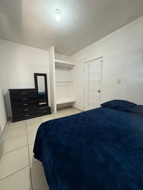 Recámara principal con cama matrimonial, edredón azul marino, cómoda oscura, espejo y clóset blanco