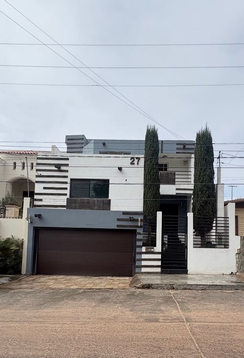 Fachada de casa moderna de dos pisos con puerta de cochera café y cipreses al frente