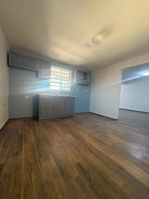 Cocina y sala con piso de madera, gabinetes grises de acero, pared acento azul y ventana