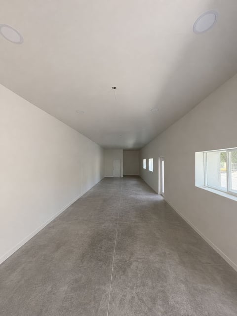 Sala moderna vacía con piso de concreto, paredes blancas, luces empotradas en techo y ventana grande al fondo
