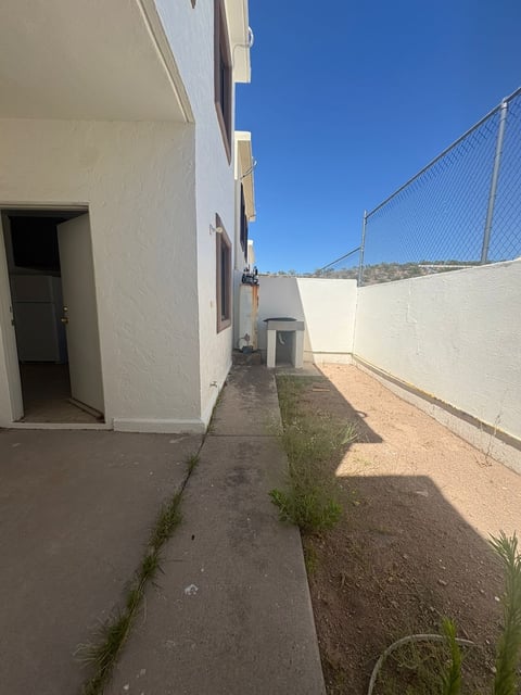 Patio lateral con piso de concreto, puerta trasera, lavadero exterior, muro blanco y barda de malla con cerro al fondo