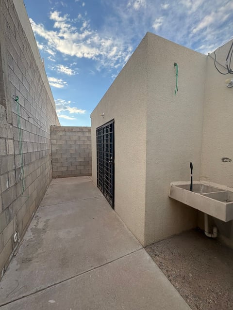 Patio trasero con piso de concreto, paredes color beige, puerta metálica y lavadero blanco bajo cielo azul