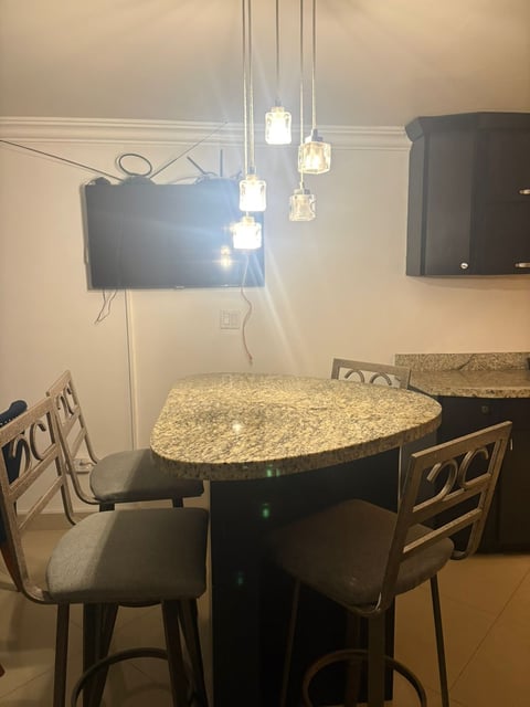Comedor con mesa ovalada de granito, dos sillas de madera y lámparas colgantes con TV montada