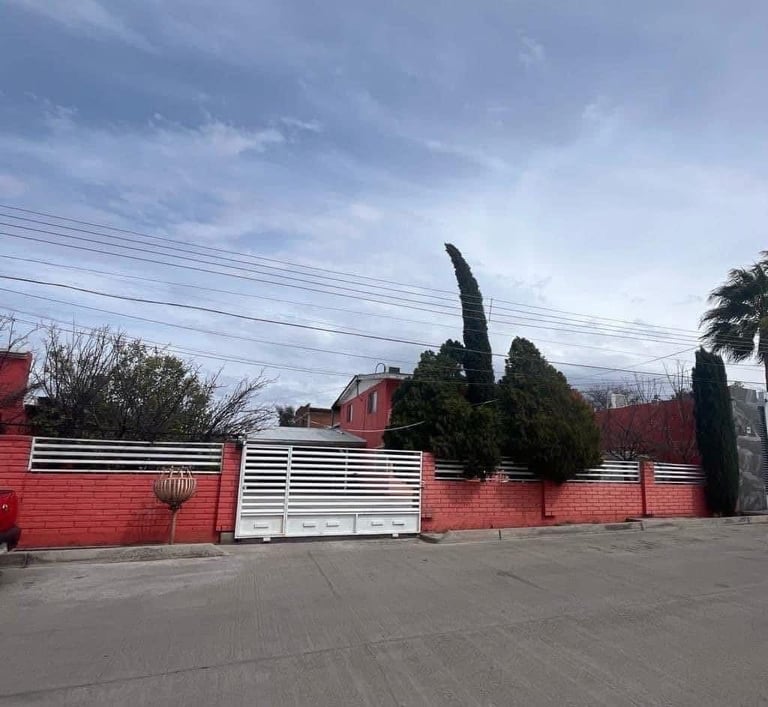 Red brick residential property with white metal gates and tall cypress trees, set against a cloudy sky with power lines visible