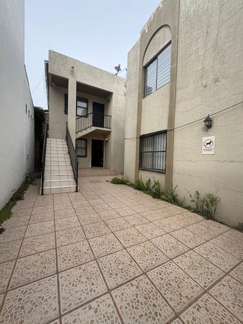 Entrada de patio de concreto a edificio residencial beige de varios pisos con escaleras, ventanas y protecciones metálicas