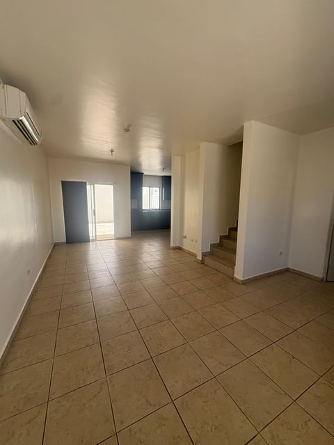 Sala abierta con piso de loseta, minisplit en techo, puerta azul corrediza, cocina al fondo y escaleras de loseta