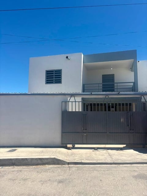 Fachada de casa blanca de dos pisos con portón metálico gris y balcón bajo cielo azul
