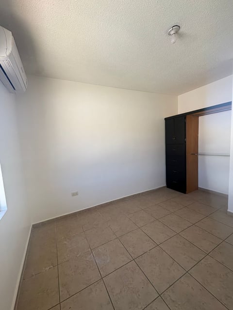 Recámara vacía con piso de loseta beige, minisplit, paredes blancas y clóset con puertas corredizas oscuras