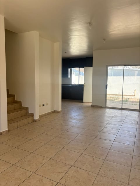 Sala abierta con piso de loseta, escaleras de madera, paredes blancas y puertas de vidrio hacia el patio