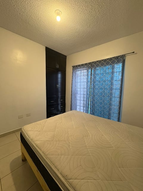 Recámara con paredes color crema, foco de techo, clóset negro, cortinas con patrón y colchón blanco al frente