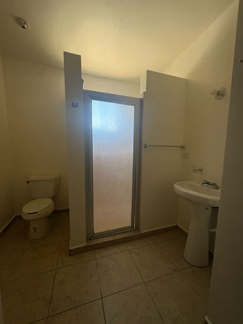 Baño con inodoro, lavamanos tipo pedestal, regadera con cancel de vidrio y piso de loseta