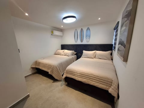 Recámara moderna con dos camas queen, cabeceras negras, ropa de cama beige, cuadros de surf y luces LED en techo