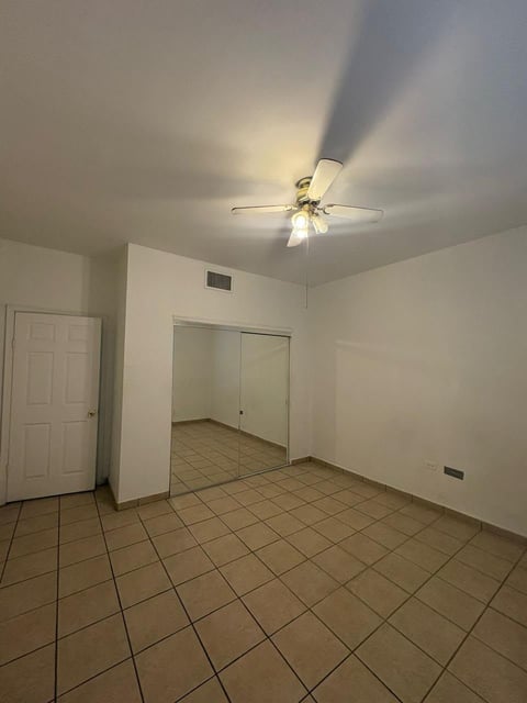 Recámara vacía con piso de loseta, paredes blancas, ventilador de techo con luces, puerta blanca a la izquierda y acceso a otra habitación