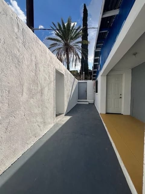 Pasillo exterior angosto entre edificios blancos con palmeras y cipreses hacia una puerta blanca