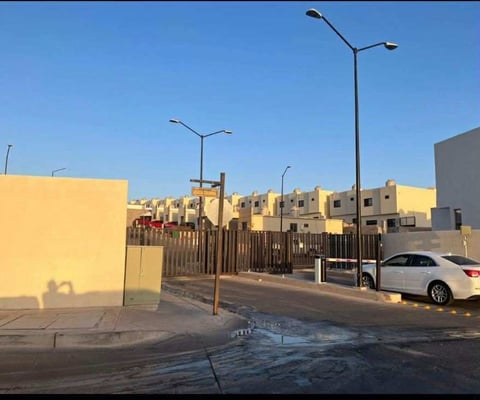 Vista del fraccionamiento con casas tipo townhouse color amarillo, lámpara de calle, auto estacionado y barda metálica bajo cielo despejado