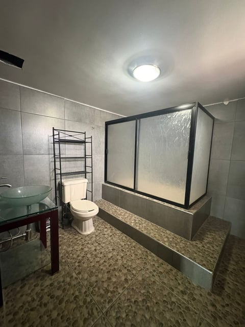 Baño con muros de loseta blanca, regadera de vidrio y repisas metálicas