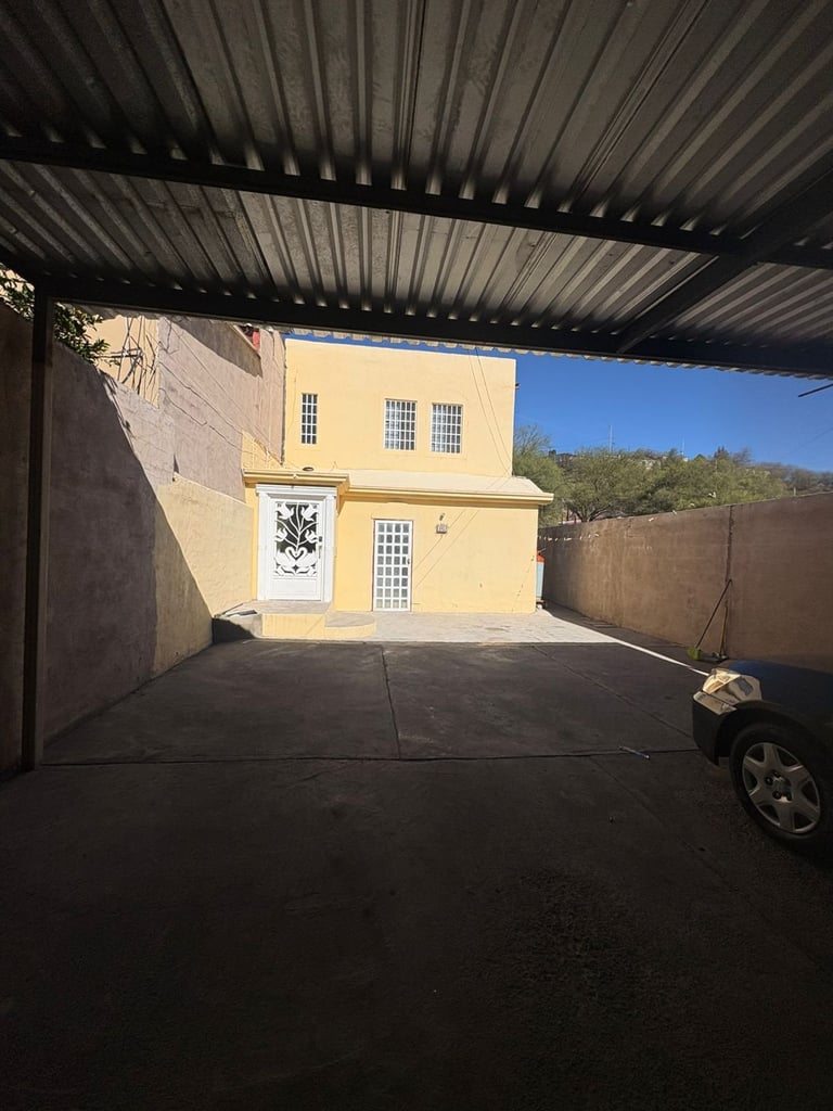 Yellow house with white-trimmed windows viewed from under a metal carport, with a parked car and desert hills visible