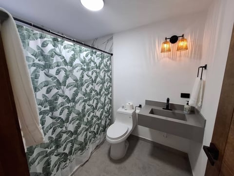 Baño moderno con lavabo gris, inodoro blanco, cortina de regadera con hojas tropicales y luces colgantes de latón