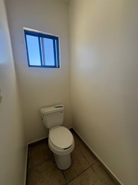 Baño con inodoro blanco, paredes beige, piso de loseta y ventana con marco azul cerca del techo
