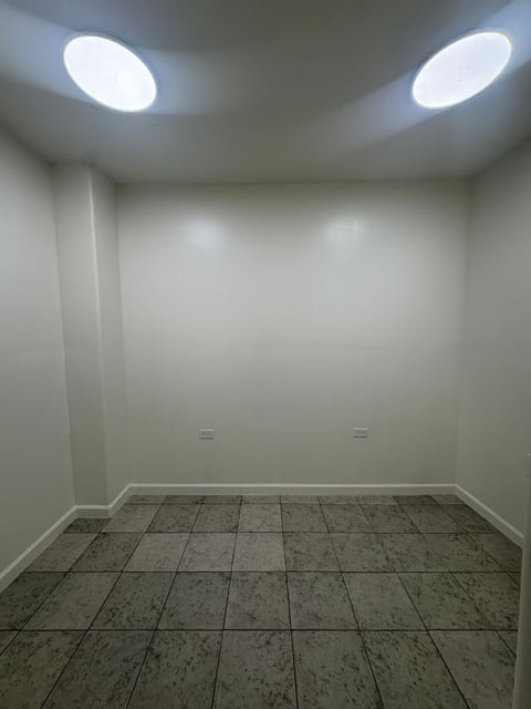 Habitación vacía con paredes blancas, piso de loseta gris y dos focos ovalados en el techo