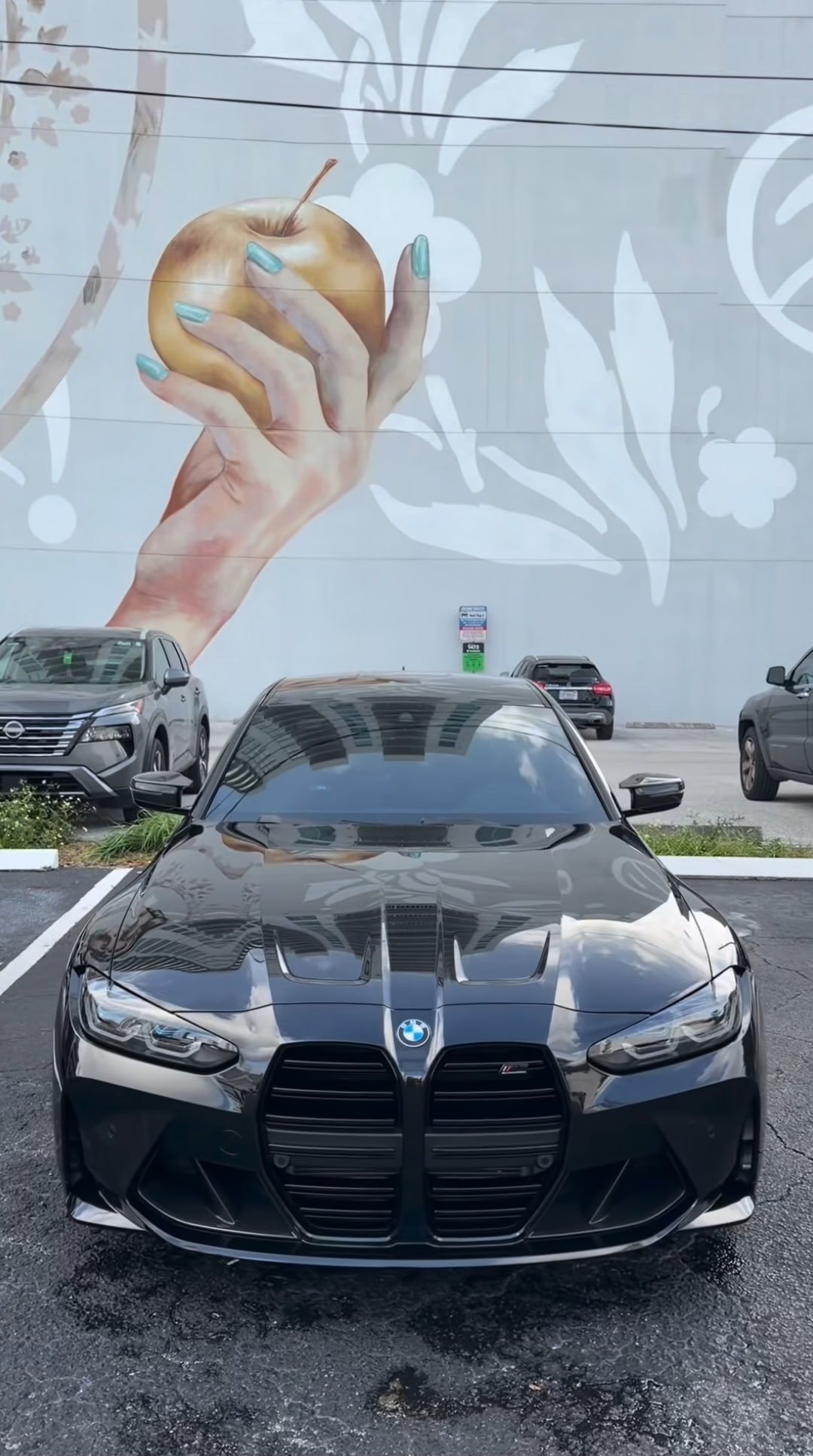 Black BMW sports car parked in front of a mural depicting a hand holding a golden apple with floral designs