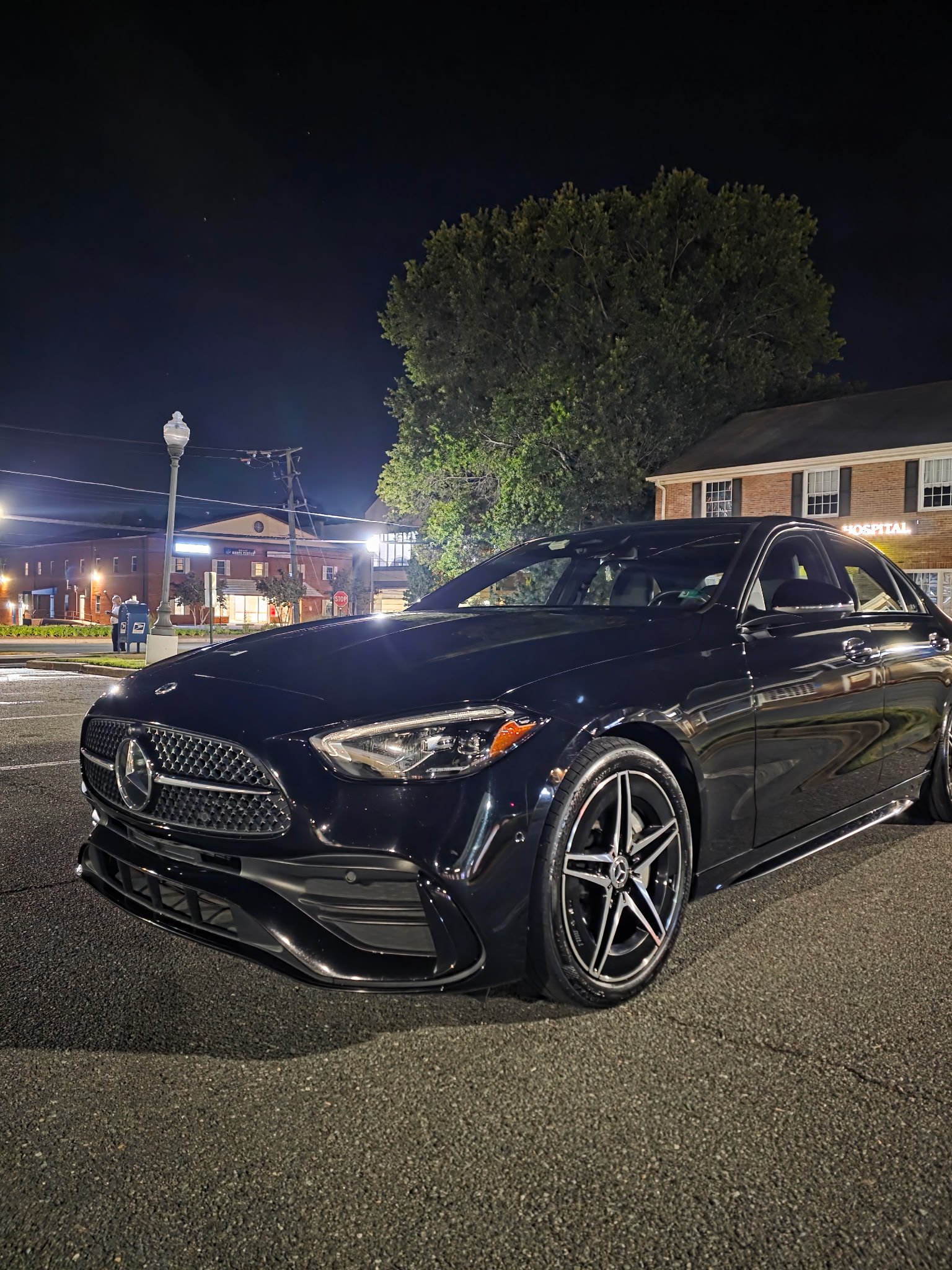 Black Mercedes-Benz luxury sedan parked on a residential street at night with illuminated buildings and tree in background
