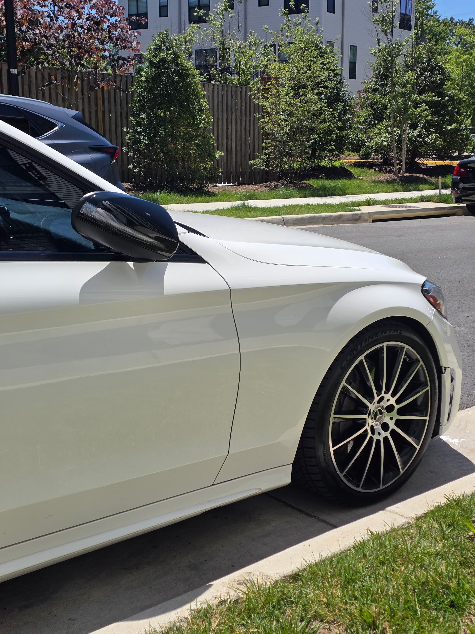 White luxury sedan parked on street with silver alloy wheel visible, residential building and landscaping in background
