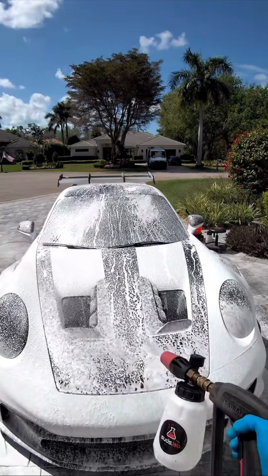White sports car being washed with a pressure washer in a residential driveway with palm trees visible