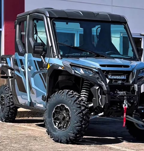 Blue and black Kawasaki utility vehicle with open cabin doors, rugged all-terrain tires, and black bumper parked outdoors