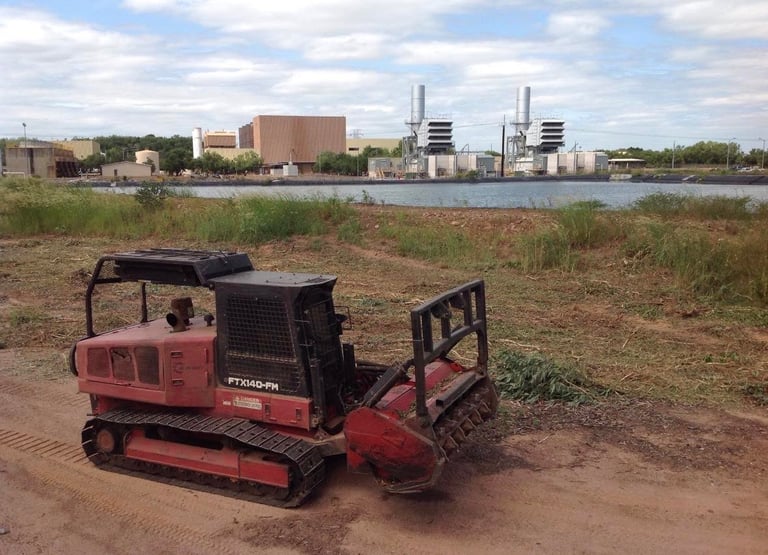 Red tracked demolition machine parked on dirt overlooking industrial waterfront with factory buildings