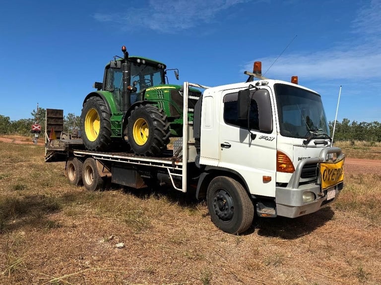 White flatbed truck transporting a green John Deere tractor on rural dirt road under blue sky