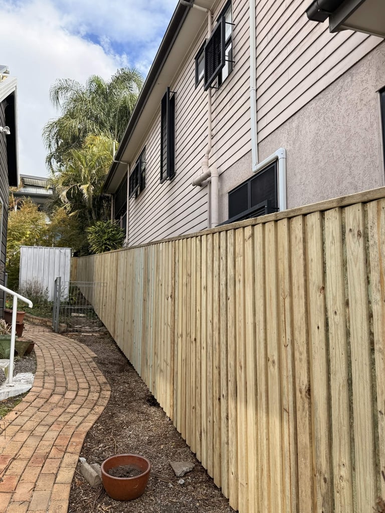 Wooden fence and brick pathway beside a residential house with corrugated metal siding and windows