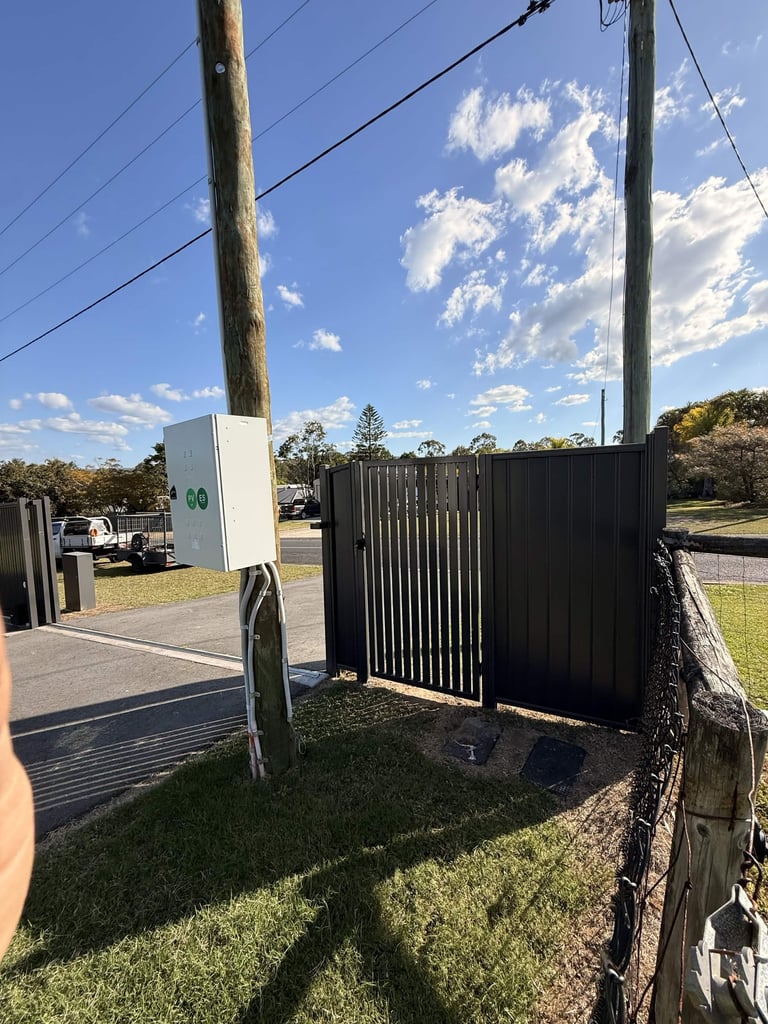 Black metal driveway gate with white EV charging station mounted on wooden pole in rural setting with clear blue sky