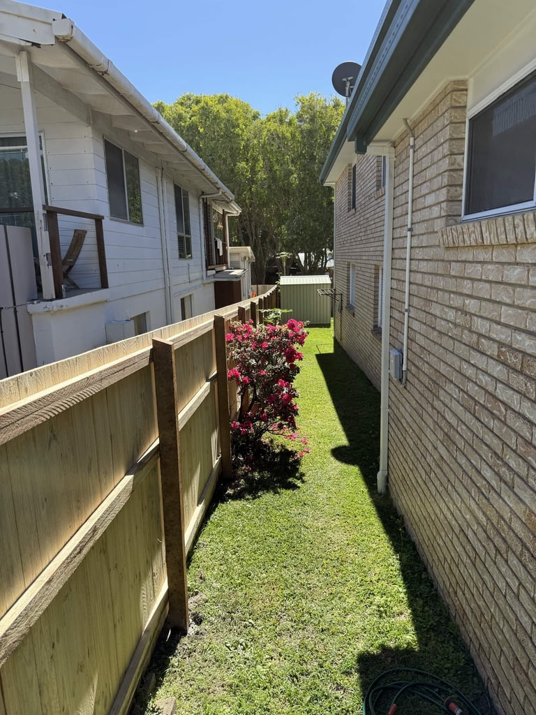Narrow side yard between two houses with wooden fence, flowering shrub, and green lawn in sunny residential area