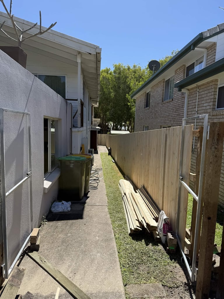 Narrow residential side yard between two houses with concrete pathway, green bin, wooden fence, and construction materials stacked against fence