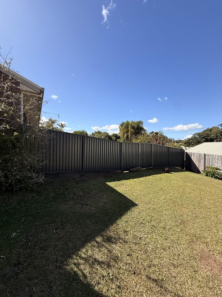 Backyard with manicured lawn, dark metal corrugated fence, trees, and clear blue sky on a sunny day