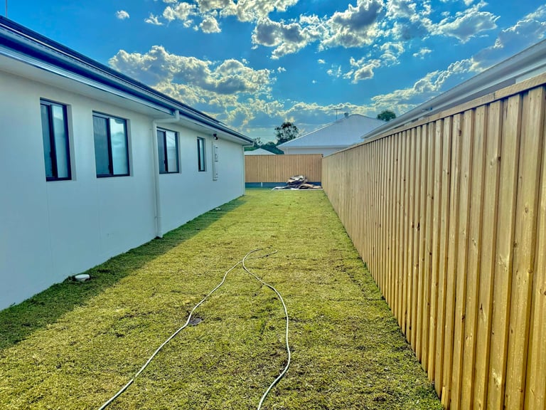 Narrow side yard between white residential building and corrugated metal fence on grassy ground under blue sky