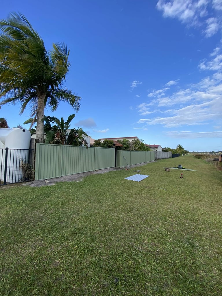 Residential property with green corrugated metal fence, palm tree, and spacious lawn under clear blue sky