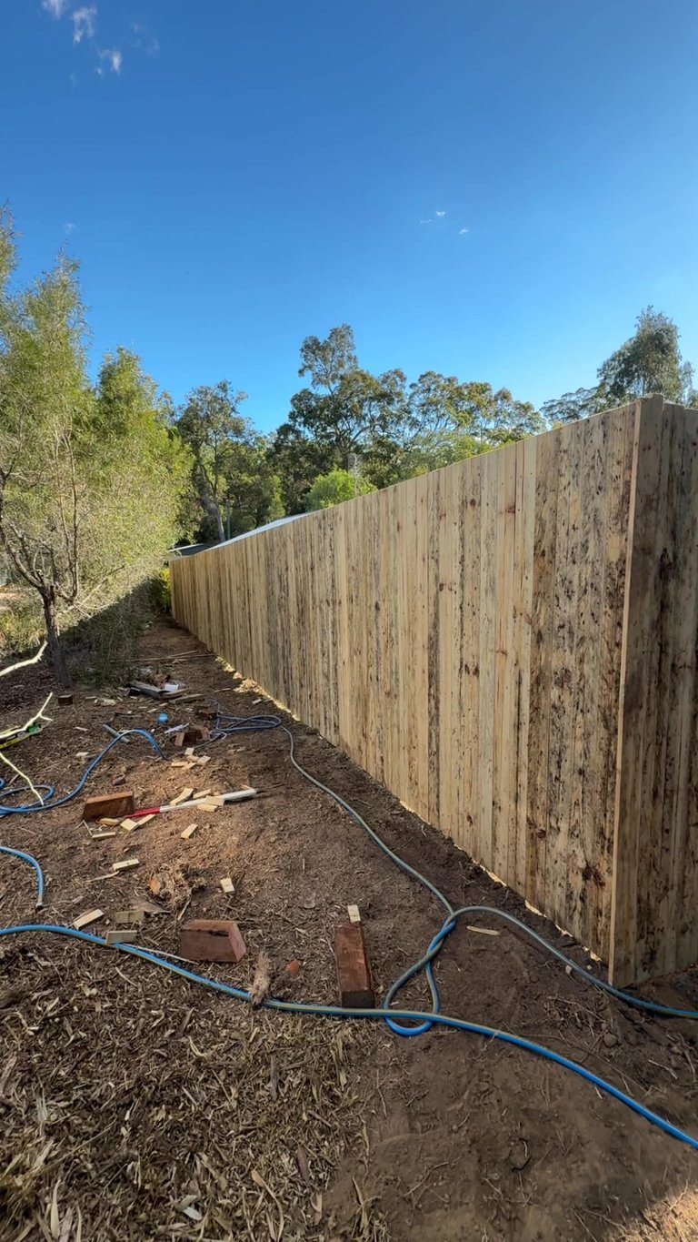 Wooden fence running along a dirt path with blue hose and trees visible under clear blue sky