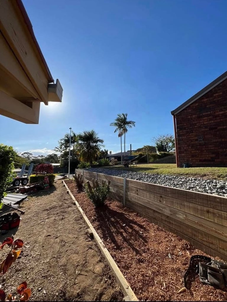 Residential backyard with wooden fence, garden beds with mulch, palm trees, and clear blue sky