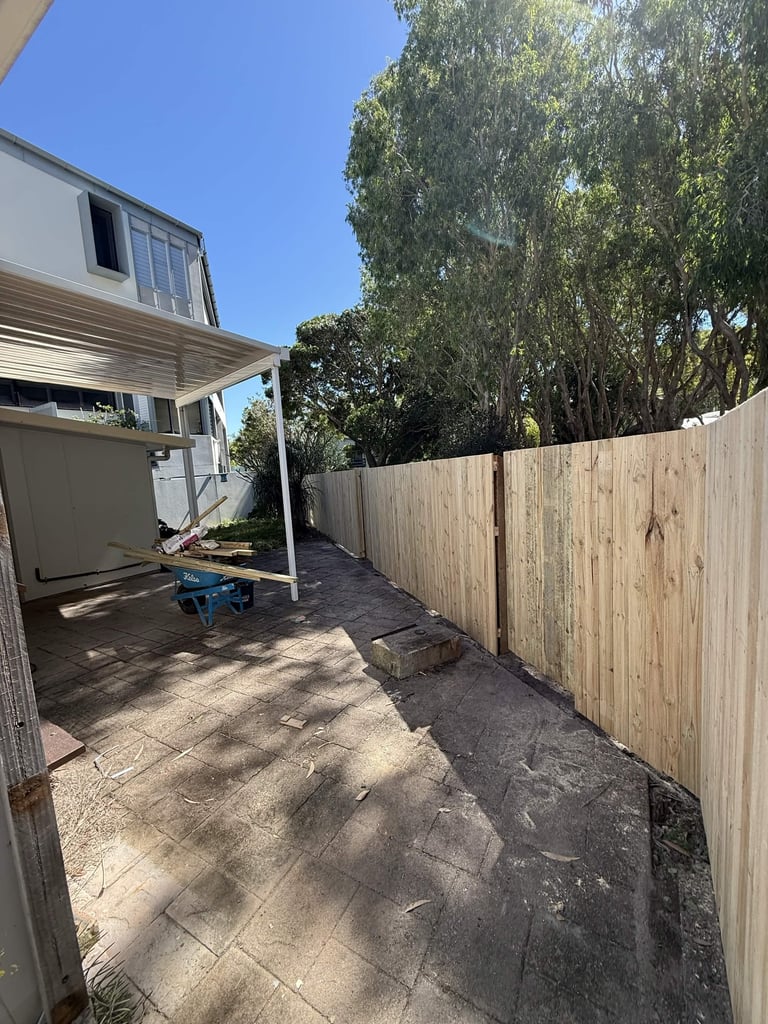 Backyard patio area with concrete flooring, wooden fence, pergola structure, and tall eucalyptus trees under blue sky