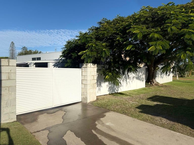 White corrugated metal driveway gate with concrete posts next to a house, surrounded by green landscaping and grass